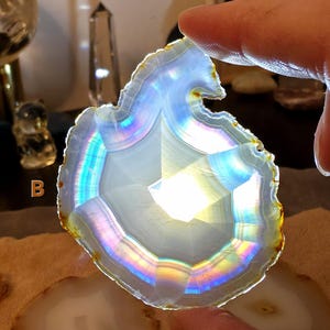 May include: A translucent, iridescent agate slice with a white centre and a rainbow-coloured edge. The stone is held by a person's fingers, showcasing its natural patterns and colours. The agate has a polished surface and a unique, organic shape.