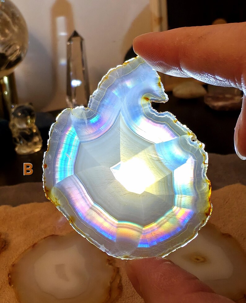 May include: A translucent, iridescent agate slice with a white centre and a rainbow-coloured edge. The stone is held by a person's fingers, showcasing its natural patterns and colours. The agate has a polished surface and a unique, organic shape.