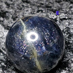 May include: A dark blue crystal sphere with a shimmering, speckled appearance. The sphere has a polished surface with a light-colored vein running through it. The crystal ball is a decorative item.
