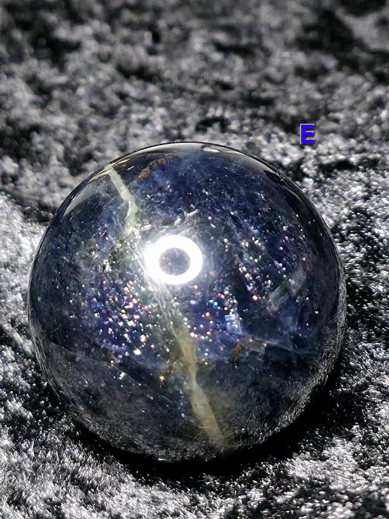 May include: A dark blue crystal sphere with a shimmering, speckled appearance. The sphere has a polished surface with a light-colored vein running through it. The crystal ball is a decorative item.