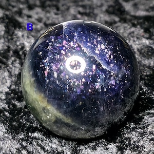 May include: A polished, spherical gemstone with a deep blue and purple color scheme. The sphere has a speckled, glittery appearance, with hints of green and white. The letter "B" is visible in the upper left corner. The gemstone is likely used for decorative purposes.