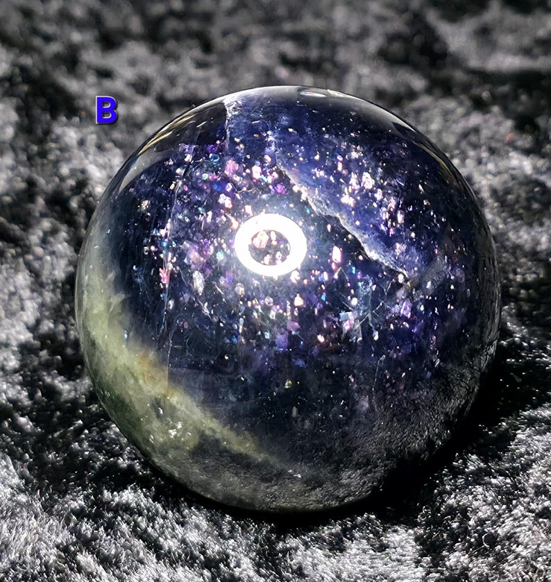 May include: A polished, spherical gemstone with a deep blue and purple color scheme. The sphere has a speckled, glittery appearance, with hints of green and white. The letter "B" is visible in the upper left corner. The gemstone is likely used for decorative purposes.
