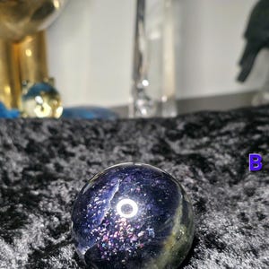 May include: A dark blue and green crystal sphere with a speckled, iridescent pattern. The sphere has a polished surface and a circular design. The background includes other crystals and decorative objects.