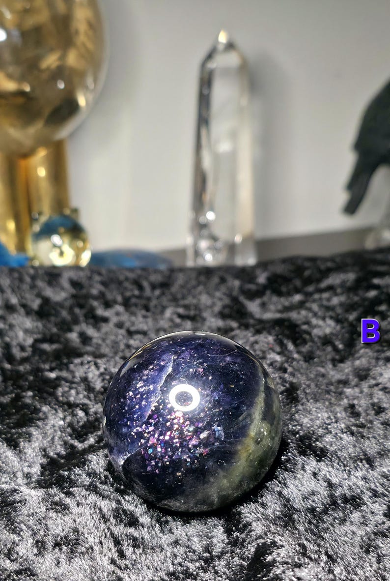 May include: A dark blue and green crystal sphere with a speckled, iridescent pattern. The sphere has a polished surface and a circular design. The background includes other crystals and decorative objects.