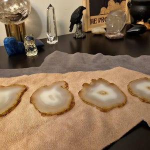 May include: Four agate coasters with a natural, organic shape. Each coaster features a white centre surrounded by a band of light brown and a darker brown edge. They are displayed on a tan cloth.