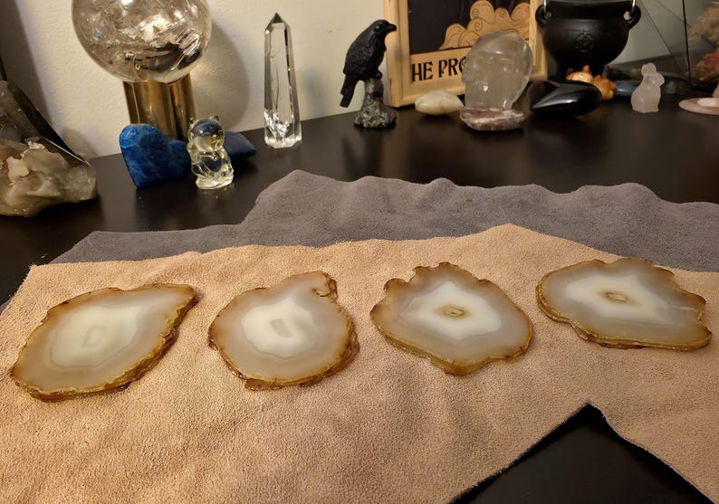 May include: Four agate coasters with a natural, organic shape. Each coaster features a white centre surrounded by a band of light brown and a darker brown edge. They are displayed on a tan cloth.