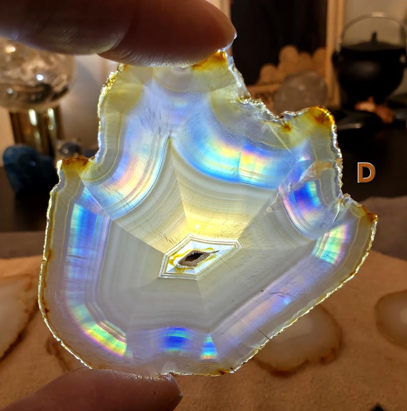 May include: A polished agate slice with a natural, uneven edge. The stone exhibits a captivating display of colours, including iridescent blues, yellows, and greens. The centre features a small, dark, diamond-shaped inclusion. The letter "D" is visible in the background.
