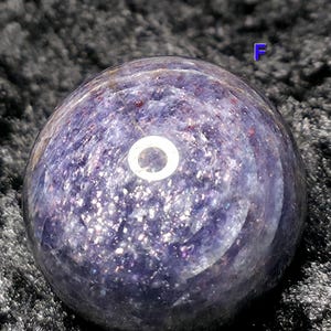 May include: A polished, spherical gemstone in shades of purple and lavender, speckled with iridescent inclusions. The orb has a smooth, reflective surface, with a bright, circular highlight. The letter "F" is visible in the background.