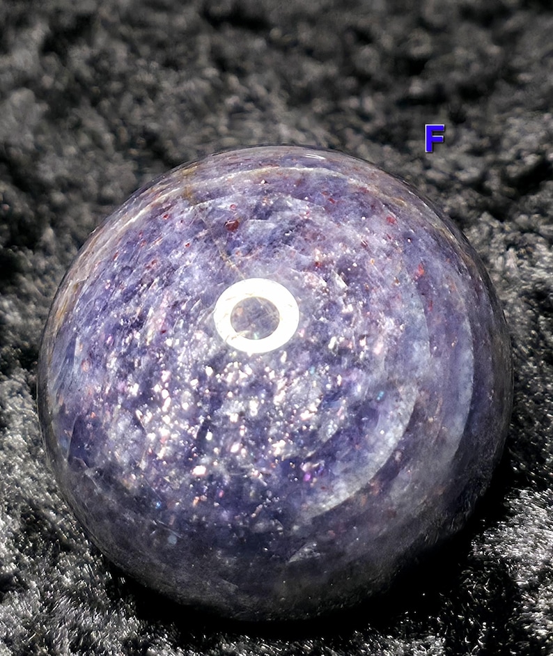 May include: A polished, spherical gemstone in shades of purple and lavender, speckled with iridescent inclusions. The orb has a smooth, reflective surface, with a bright, circular highlight. The letter "F" is visible in the background.