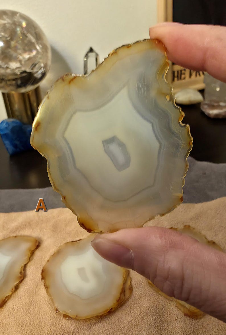 May include: A polished agate slice, displaying a pattern of white, grey, and brown concentric bands. The natural stone has an irregular shape and a translucent quality. Other agate slices are visible below. The image showcases natural stone decor.