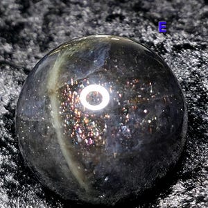 May include: A dark, iridescent sphere with a polished surface. The sphere has a mix of colors, including black, silver, and hints of gold and purple. The sphere is round and appears to be made of a gemstone or mineral.