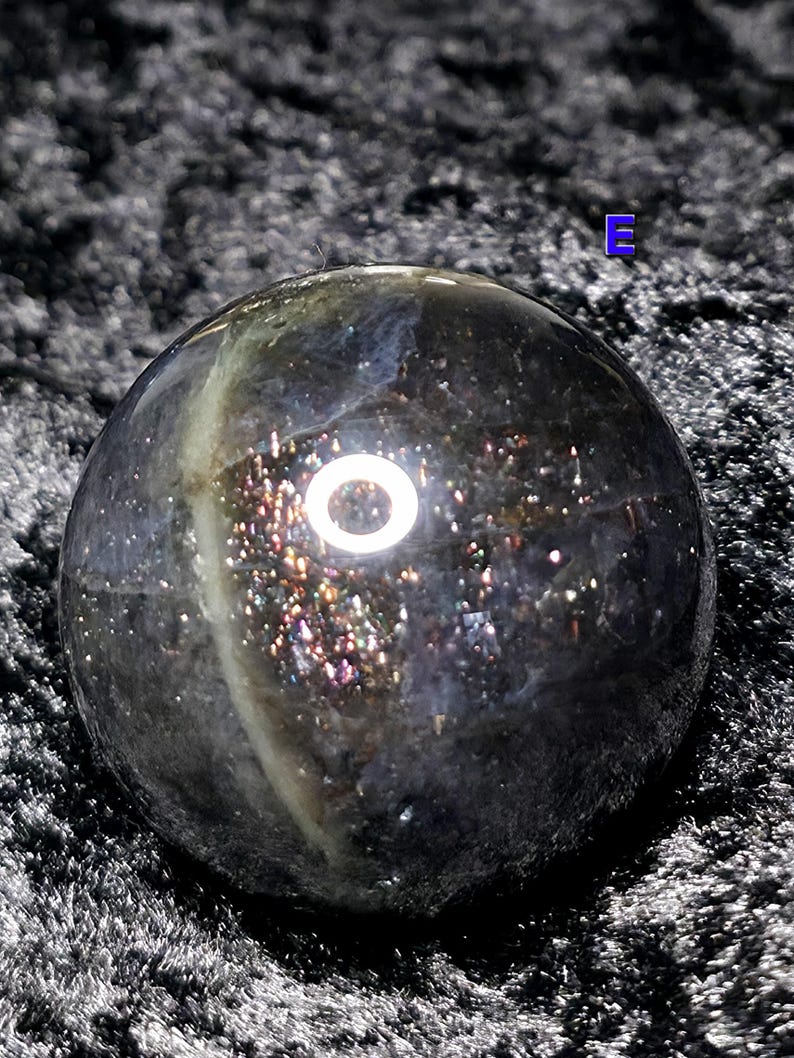 May include: A dark, iridescent sphere with a polished surface. The sphere has a mix of colors, including black, silver, and hints of gold and purple. The sphere is round and appears to be made of a gemstone or mineral.