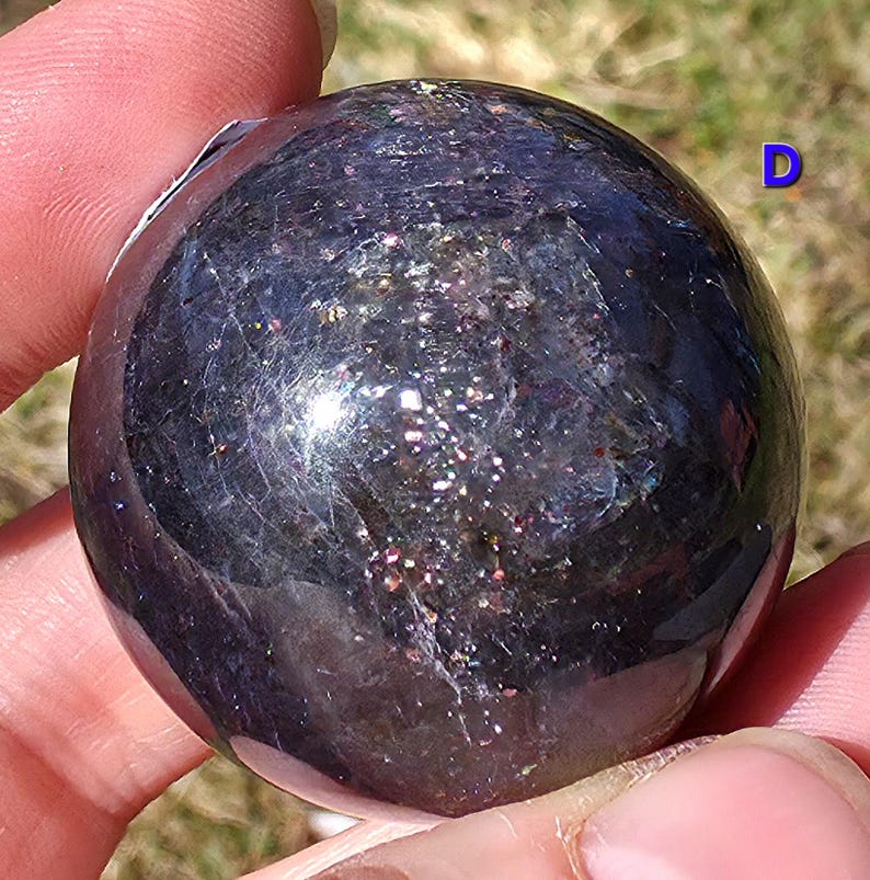 May include: A dark, iridescent sphere with a polished surface. The sphere has a speckled appearance with hints of pink, purple, and gold. The letter "D" is visible in the upper right corner. The sphere is held in a hand.