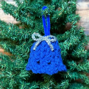 May include: A blue crocheted bell Christmas ornament with a silver bow and blue velvet hanger, displayed on a green artificial Christmas tree. The bell has a textured pattern, a festive holiday decoration.