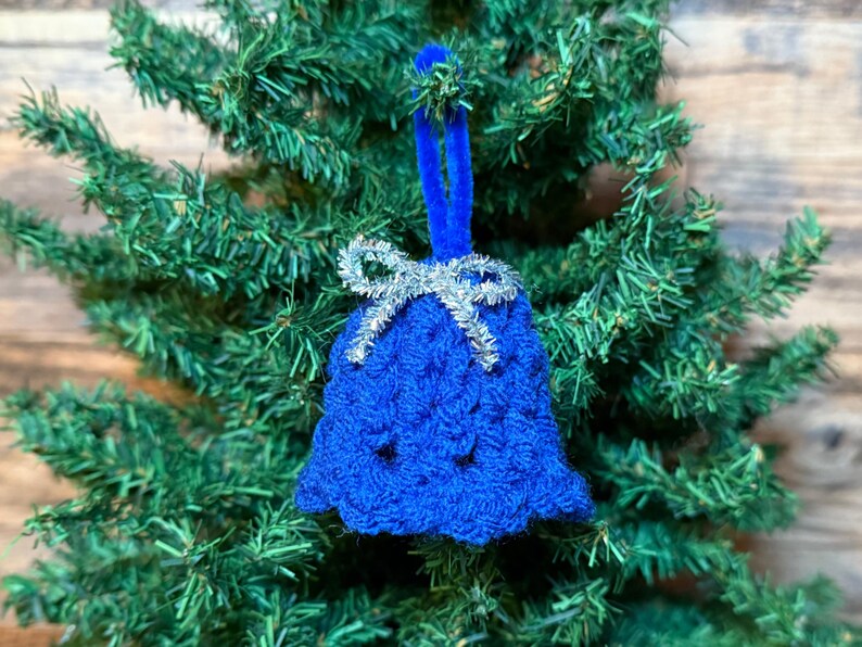 May include: A blue crocheted bell Christmas ornament with a silver bow and blue velvet hanger, displayed on a green artificial Christmas tree. The bell has a textured pattern, a festive holiday decoration.
