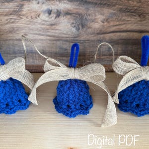 May include: Three blue crocheted bell ornaments with tan burlap bows and blue loops for hanging. The bells are arranged on a light brown wooden surface with a darker wooden background. The text "Digital PDF" is visible.