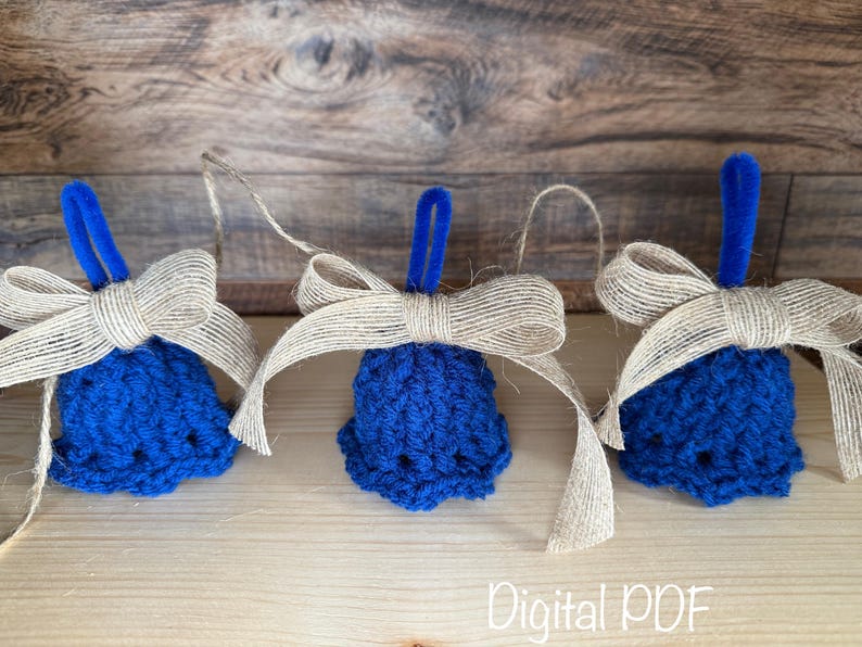 May include: Three blue crocheted bell ornaments with tan burlap bows and blue loops for hanging. The bells are arranged on a light brown wooden surface with a darker wooden background. The text "Digital PDF" is visible.