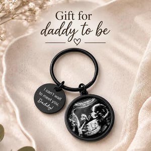 Könnte beinhalten: Ein schwarzer Metallschlüsselanhänger mit zwei runden Anhängern. Einer zeigt ein schwarz-weißes Ultraschallbild eines Fötus, der andere ist mit dem Text graviert: I can't wait to meet you, Daddy! Der Text Gift for daddy to be ist sichtbar.