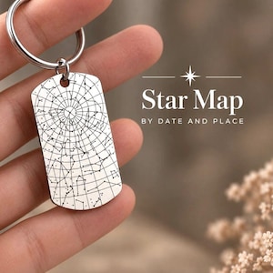 Könnte beinhalten: Ein silberfarbener Metall-Schlüsselanhänger in Form einer Erkennungsmarke mit einer schwarz gravierten Sternenkarte, kreisförmigen Gitterlinien und Sternbildern. Der Text "Star Map BY DATE AND PLACE" ist in Weiß mit einem Sternsymbol gedruckt.