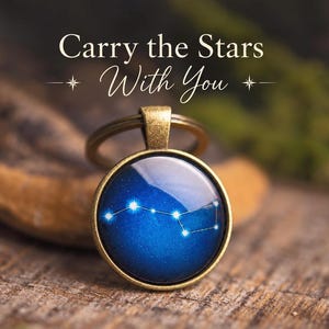 Op de afbeelding: Een ronde sleutelhanger met een antiek bronskleurig metalen frame en een diepblauwe ronde inzet met een wit sterrenbeeld. De tekst "Carry the Stars With You" is in het wit boven de sleutelhanger gedrukt.