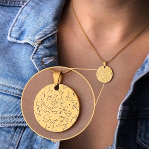 Può includere: Una collana dorata con un ciondolo rotondo che mostra un design di costellazione dettagliato. La collana è indossata sopra una giacca di jeans. Il ciondolo ha un diametro di circa 2,5 cm.