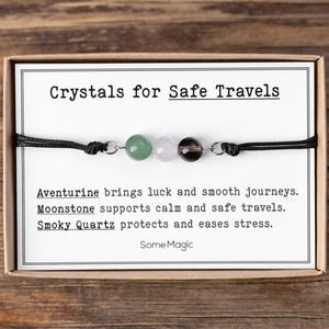 Puede incluir: Un brazalete con tres cuentas de piedras preciosas: aventurina verde, piedra lunar rosa y cuarzo ahumado. El brazalete está en una tarjeta que dice "Crystals for Safe Travels". La tarjeta también enumera las propiedades de cada cristal.