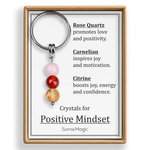 May include: A keychain with a silver ring and three gemstone beads: rose quartz, carnelian, and citrine. The card reads "Crystals for Positive Mindset" and lists the benefits of each crystal. The keychain is in a wooden frame.