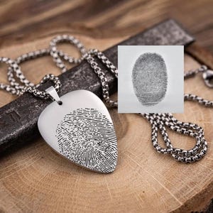 Op de afbeelding: Een zilverkleurige hangerketting met een hartvormige hanger met een vingerafdrukontwerp. De ketting heeft een ketting met een vierkant schakelpatroon. Een kleine afbeelding van een vingerafdruk staat op de achtergrond.