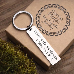 May include: A silver rectangular keychain with the text "Drive safe handsome. I love you -K" and a heart symbol. The keychain is attached to a silver ring. The keychain rests on a brown box with the words "Some Magic handmade Jewelry".