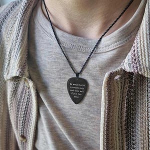 Pode incluir: Colar com pingente de palheta de guitarra preta com o texto "We would build a rocket ship and then we'd fly it far away". O pingente pende de uma corrente preta. A pessoa está vestindo uma camiseta cinza e um cardigã listrado.