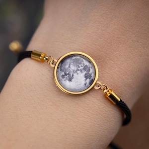 Può includere: Braccialetto dorato con un pendente circolare raffigurante un'immagine dettagliata della luna. Il braccialetto ha un cinturino in corda nera con dettagli dorati. L'immagine della luna è in tonalità di grigio e bianco.