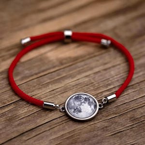 Puede incluir: Una pulsera de cordón rojo con un dije de luna plateado. El dije presenta una imagen detallada de la superficie lunar. La pulsera tiene extremos metálicos plateados ajustables.