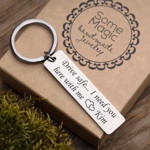 Op de afbeelding: Een zilveren rechthoekige sleutelhanger met de gegraveerde tekst "Drive safe... I need you here with me Kim" en twee hartsymbolen. De sleutelhanger is bevestigd aan een grote zilveren ring. De sleutelhanger ligt op een bruine doos met de tekst "Some Magic handmade Jewelry".