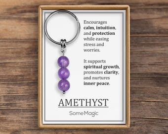 May include: A keychain with three purple amethyst beads hanging from a silver ring. The keychain is displayed on a card with text that reads "Encourages calm, intuition, and protection..." and "AMETHYST".