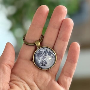 May include: A round keychain pendant featuring a detailed image of the moon in shades of gray and white. The pendant has a bronze-colored metal frame and a matching key ring.