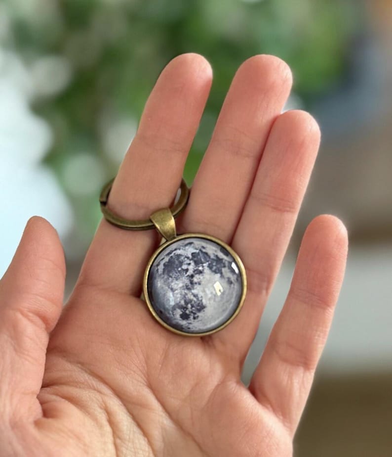 May include: A round keychain pendant featuring a detailed image of the moon in shades of gray and white. The pendant has a bronze-colored metal frame and a matching key ring.