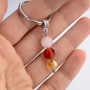 May include: A silver keychain with three gemstone beads: a light pink, a reddish-brown, and a golden-yellow. The beads are strung together and suspended from a silver ring.