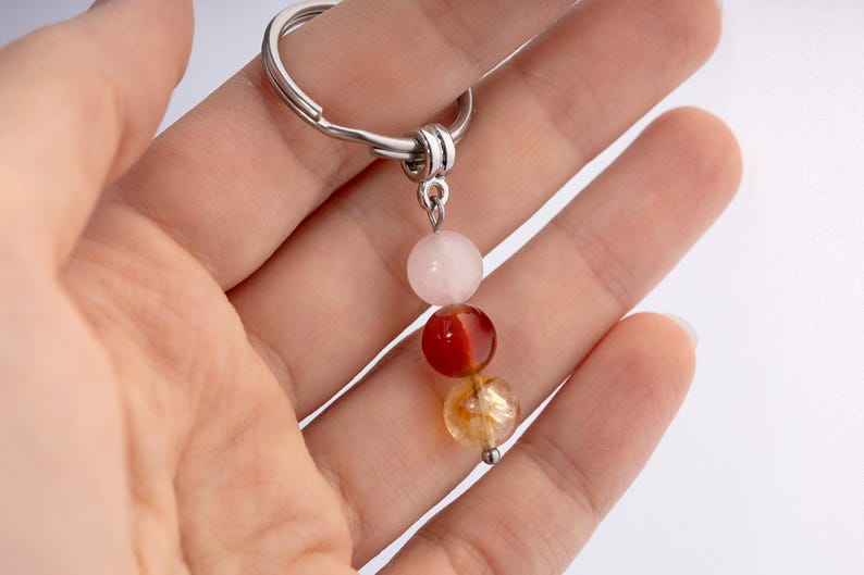 May include: A silver keychain with three gemstone beads: a light pink, a reddish-brown, and a golden-yellow. The beads are strung together and suspended from a silver ring.