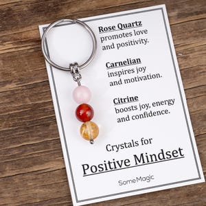 May include: A keychain with a silver ring and three gemstone beads: rose quartz, carnelian, and citrine. The accompanying card reads "Crystals for Positive Mindset" and lists the properties of each crystal.