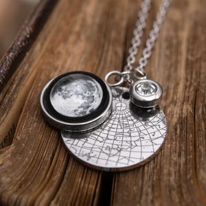 Puede incluir: Un collar plateado con colgantes de temática celestial. El collar incluye un colgante redondo con una imagen detallada de la luna, un colgante circular con un diseño de mapa estelar y un pequeño colgante de gema brillante. El collar se exhibe sobre una superficie de madera.