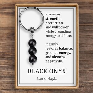 Puede incluir: Llavero de ónix negro con tres cuentas negras pulidas. La tarjeta adjunta dice: "Promueve la fuerza, la protección y la fuerza de voluntad". También menciona el equilibrio, la conexión a tierra de la energía y la absorción de la negatividad. La marca "SomeMagic".