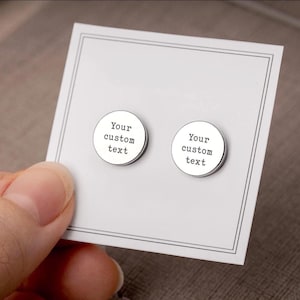 May include: A pair of silver-colored round stud earrings with the text "Your custom text" printed in black. The earrings are displayed on a white card with a black border, held by a hand.