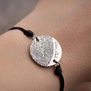 May include: A silver bracelet with a black cord. The circular pendant displays a fingerprint and the words "Love, Dad". The bracelet is worn on a wrist.