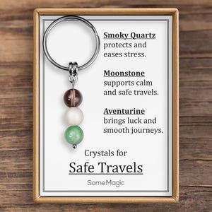 May include: A keychain with three gemstone beads: smoky quartz, moonstone, and aventurine. The keychain is attached to a card with text that reads "Crystals for Safe Travels" and descriptions of each crystal's properties. The background is a wooden surface.