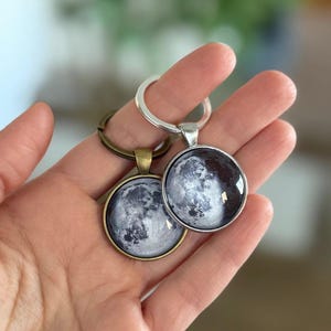 May include: Two round keychains with a moon phase design. One keychain has a silver-colored metal ring and pendant, the other has a bronze-colored ring and pendant. The moon image is a detailed grayscale photograph.