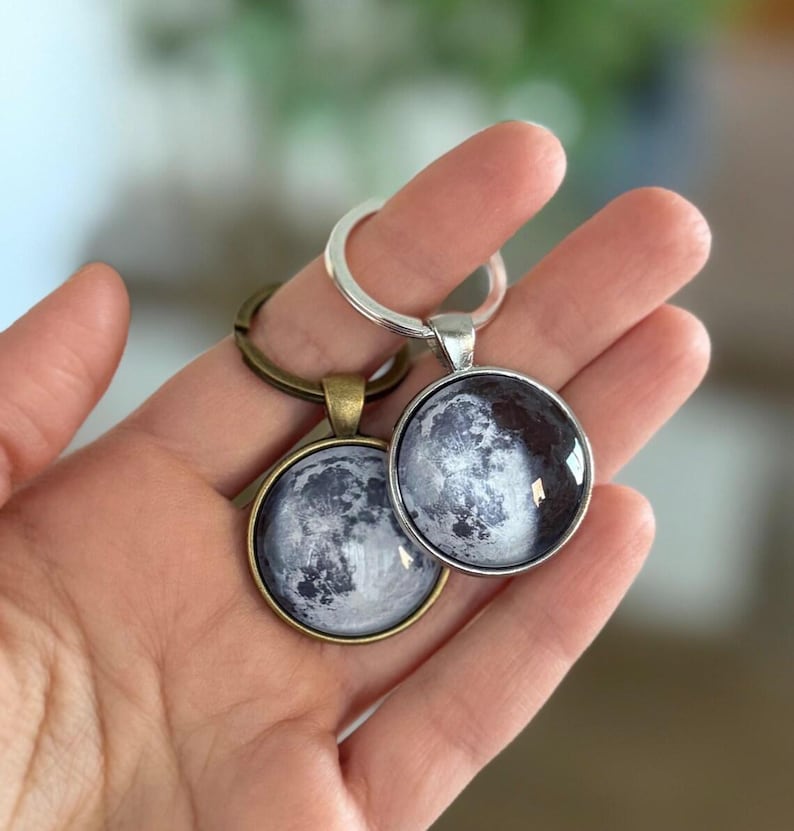 May include: Two round keychains with a moon phase design. One keychain has a silver-colored metal ring and pendant, the other has a bronze-colored ring and pendant. The moon image is a detailed grayscale photograph.