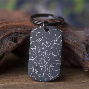 Könnte beinhalten: Ein dunkelgrauer Schlüsselanhänger in Form einer Hundemarke mit einem weißen Sternbilddesign. Der Schlüsselanhänger hat einen schwarzen Ring und wird auf einem Stück Treibholz präsentiert. Die Sternbilder sind durch weiße Linien verbunden.
