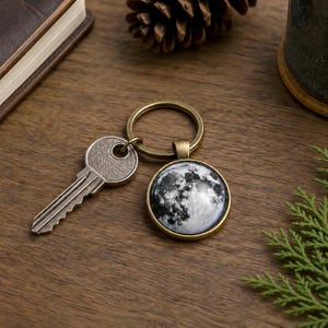 May include: A key and a keychain with a round pendant featuring a detailed image of the moon. The keychain has a brass-colored ring and frame. The key is silver. The items are on a wooden surface.