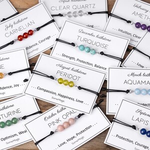 Könnte beinhalten: Eine Sammlung von Geburtssteinarmbändern mit verschiedenfarbigen Perlen, jeweils an einem schwarzen Band. Jedes Armband wird auf einer Karte mit dem Geburtsstein-Namen und -Monat präsentiert. Die Karten enthalten auch das Wort "SomeMagic".