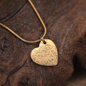 Può includere: Una collana con pendente a forma di cuore dorato. Il pendente presenta un disegno di costellazione. La catena della collana è una sottile catena a serpente dorata. La collana è esposta su una superficie di legno scuro e strutturato.